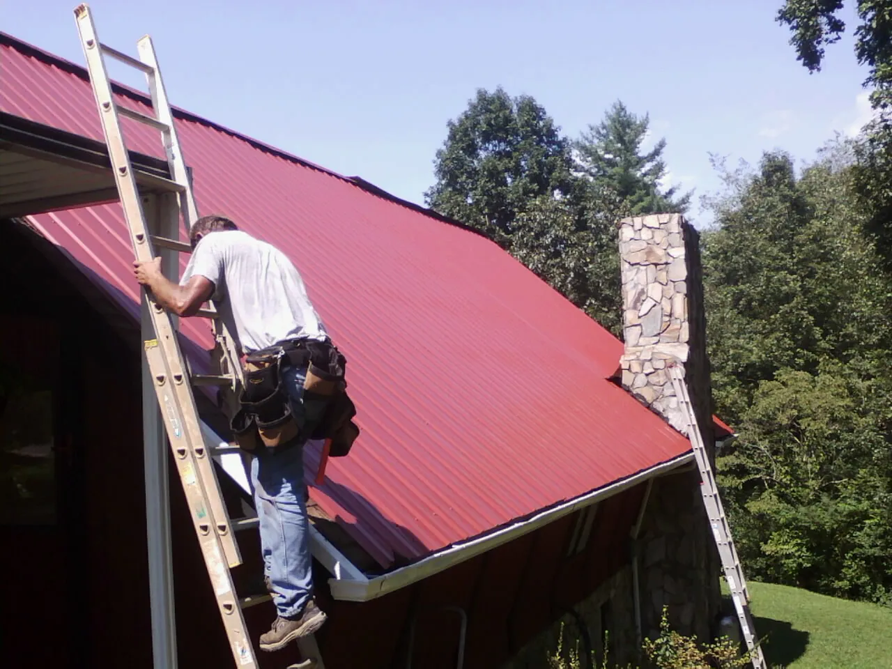 Roofer on a ladder beside a red metal roof and stone chimney — Soffit & Fascia Repair by Roofing Brothers in Tiller